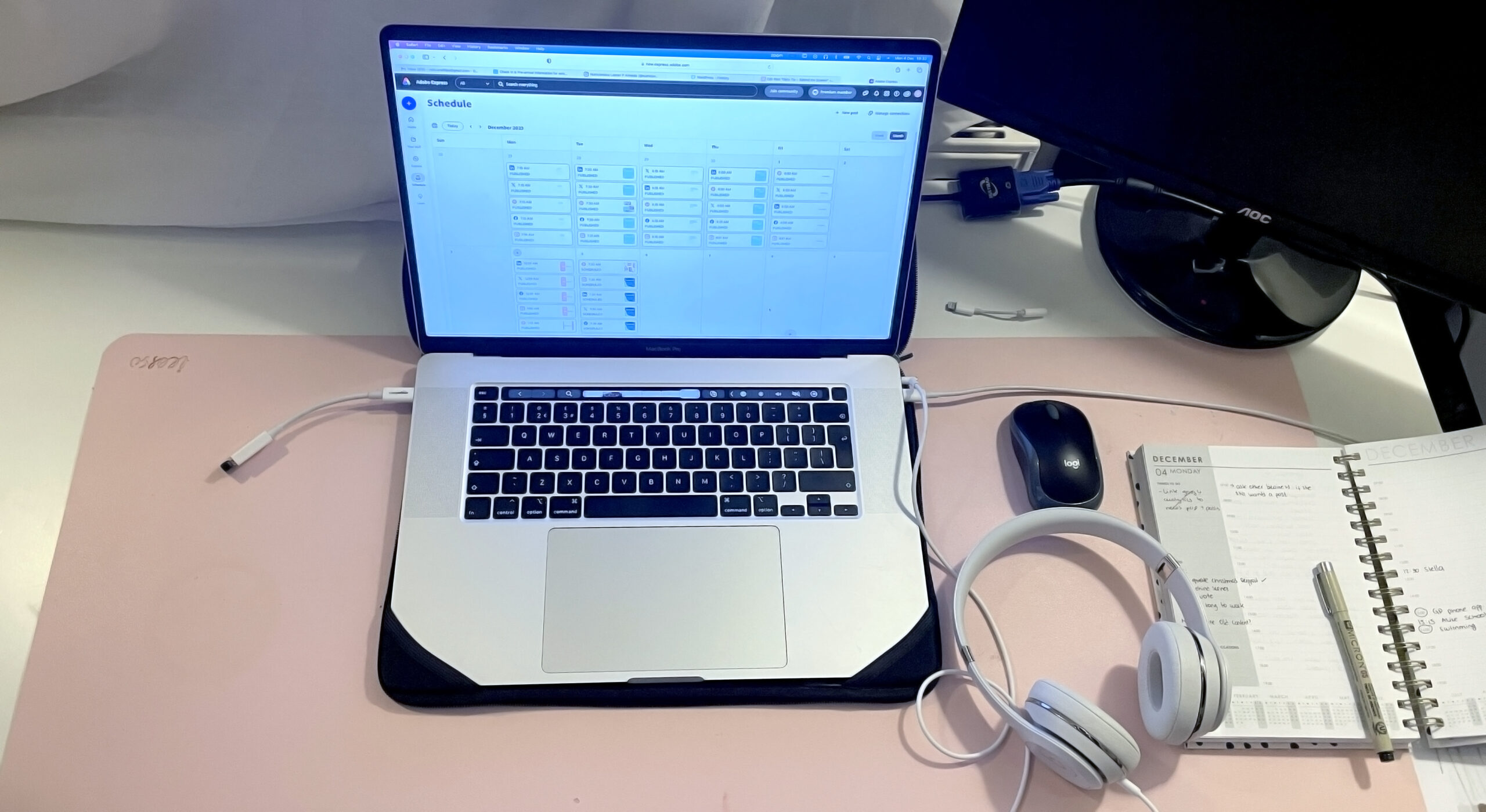 apple laptop computer opened with a schedule on top of a white desk with a pink mat underneath the computer. There's a mouse, an agenda and some headphones aswell a screen in the back