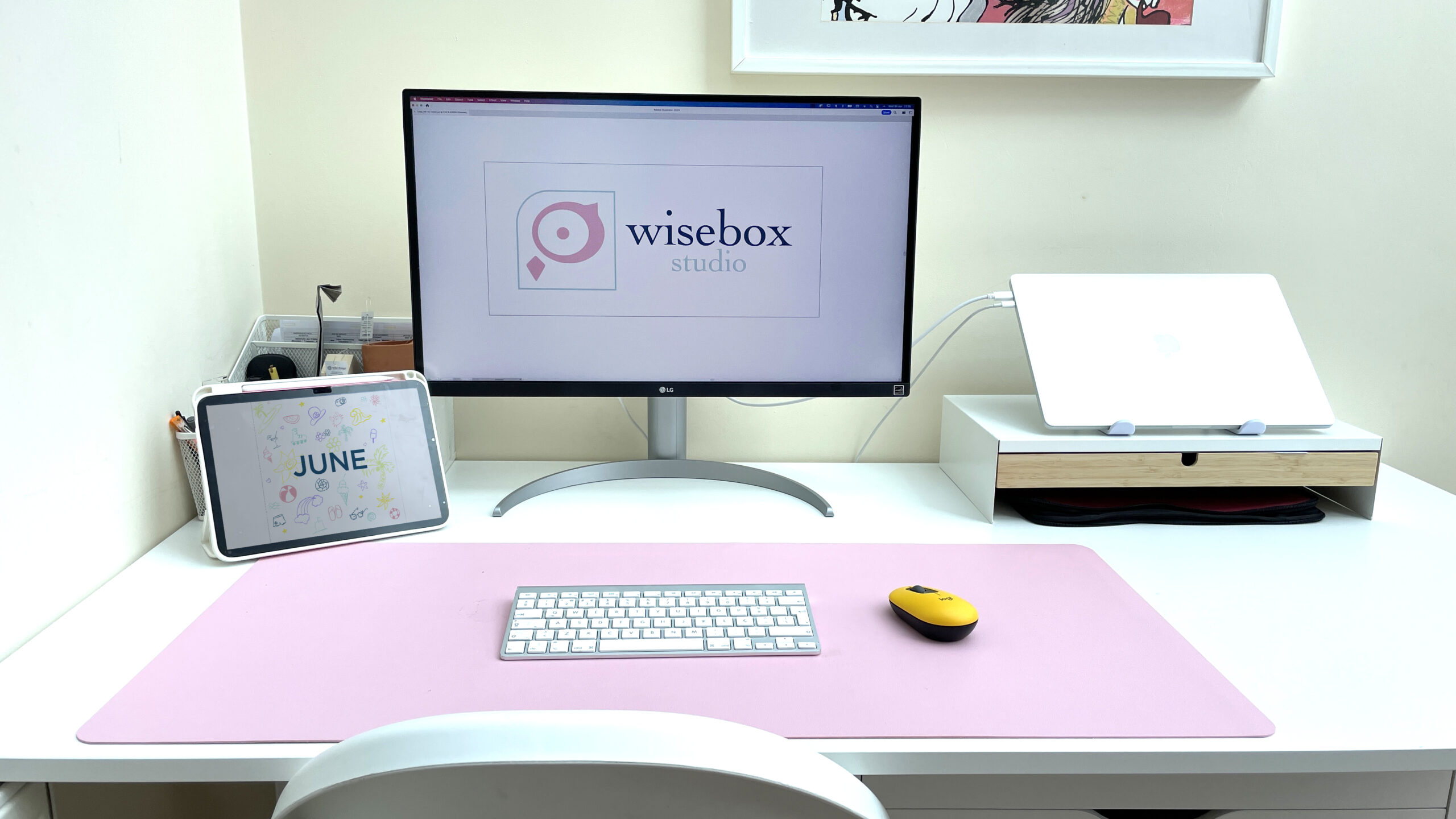 Desk with a pink pad with a monitor a laptop on a stand and an iPad with a white keyboard and a yellow mouse